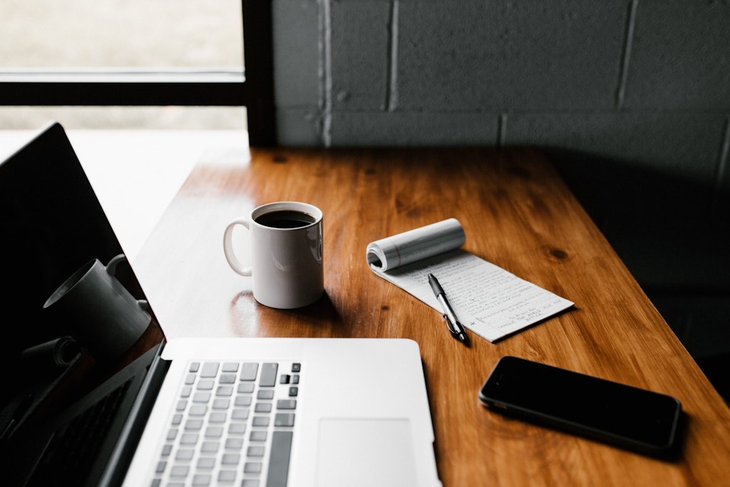 laptop, coffee cup, notebook, and cell phone on a desk